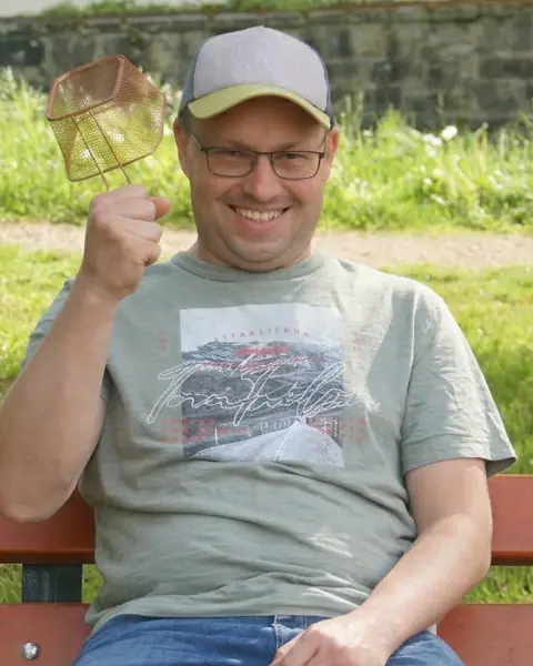 Christian wearing a cap and glasses, smiling with a raised fly swatter